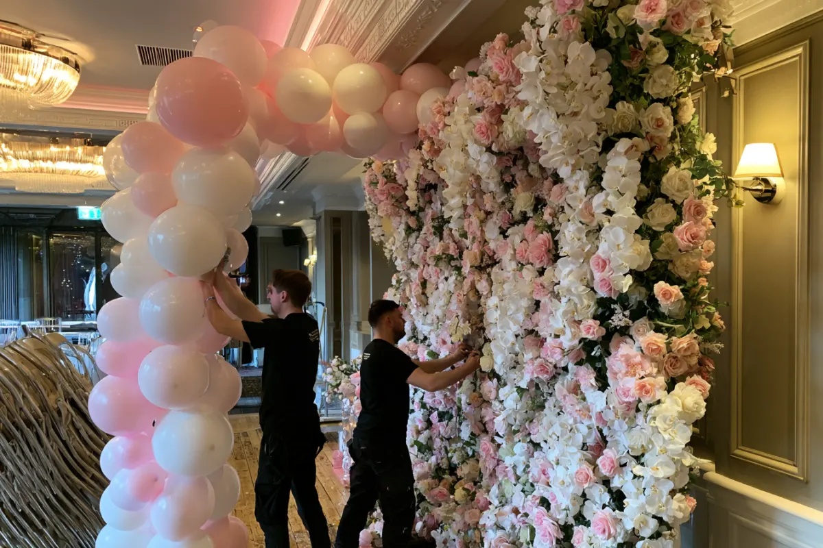 Two decorators setting up a balloon column next to a flower wall